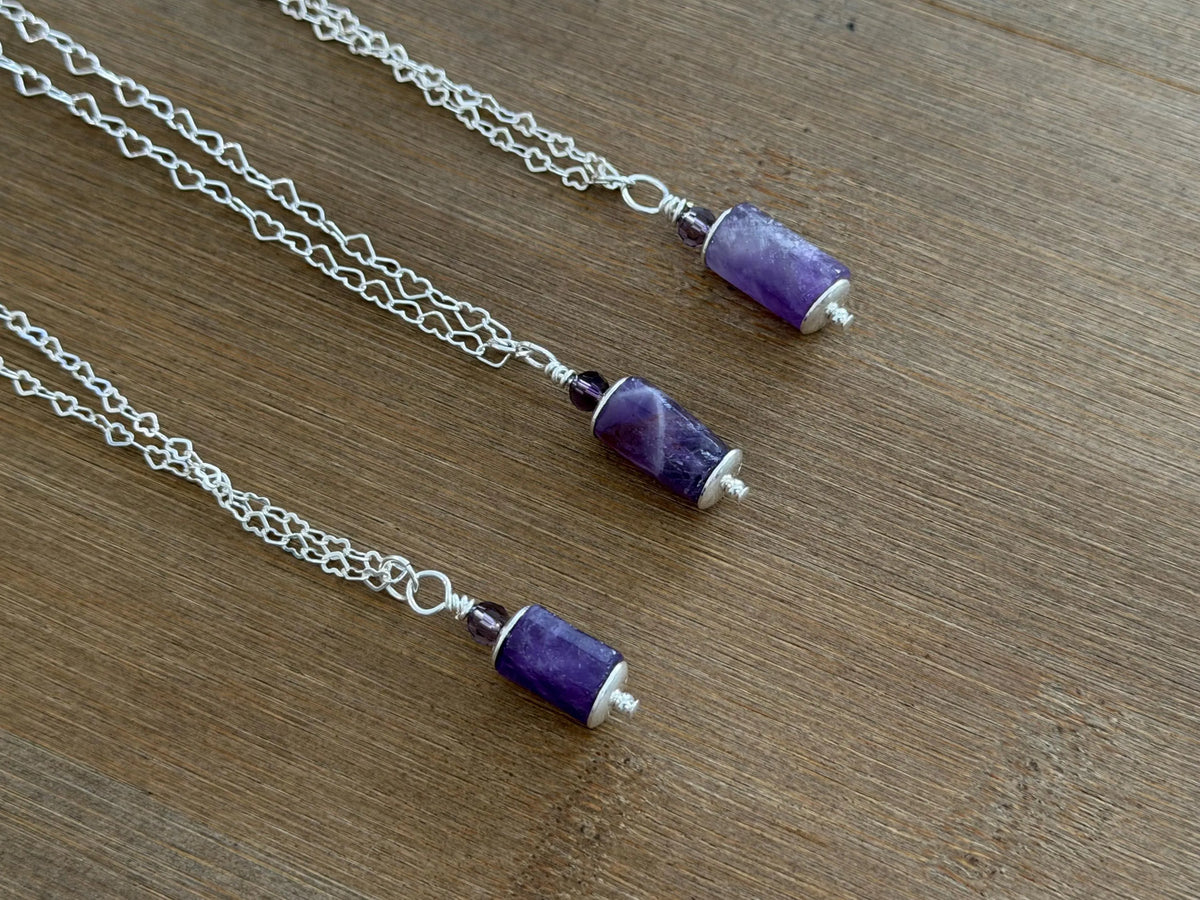 Three silver chain necklaces with cylindrical purple amethyst beads and small silver end caps, arranged diagonally on a brown wooden surface.