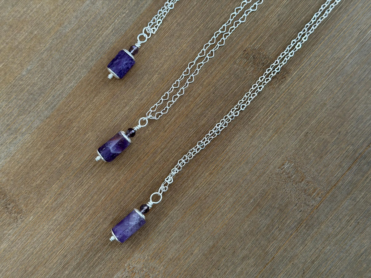 Three silver chain necklaces with cylindrical purple gemstone pendants, arranged diagonally on a wooden surface.