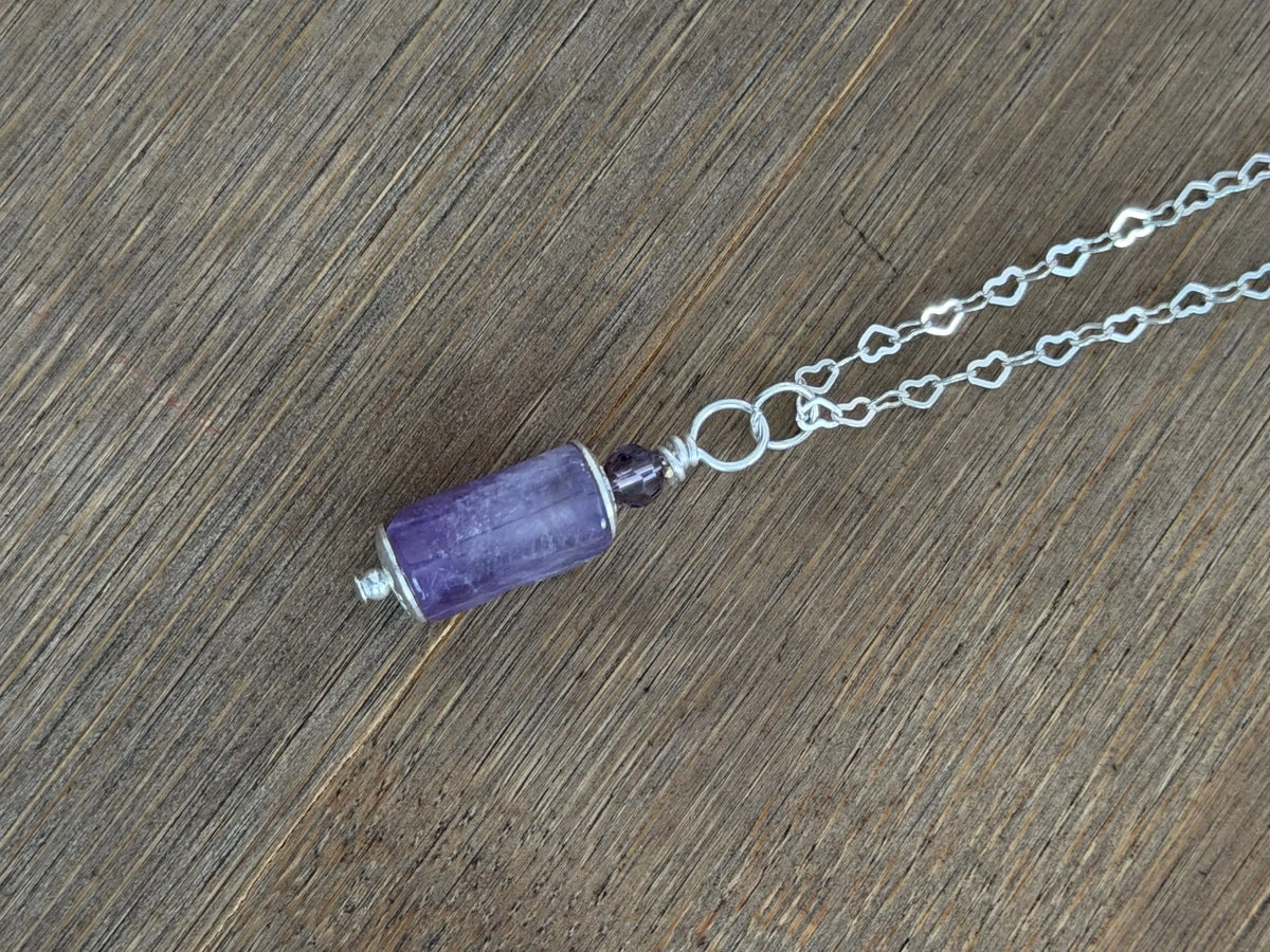 A silver chain necklace featuring a cylindrical purple stone bead pendant and a small dark accent bead, displayed diagonally on a brown wood grain surface.