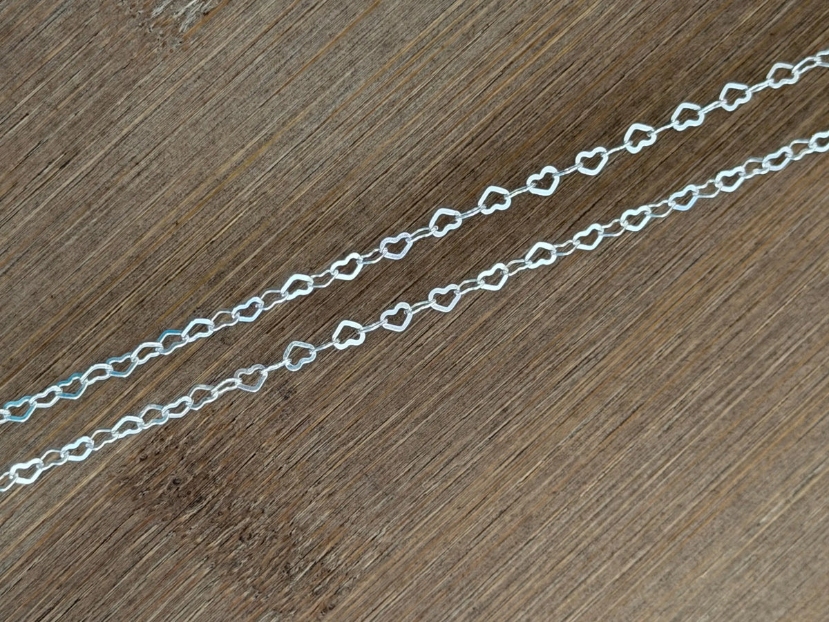 A close-up of a silver chain made of repeating open heart links, laid diagonally across a brown wood grain surface.