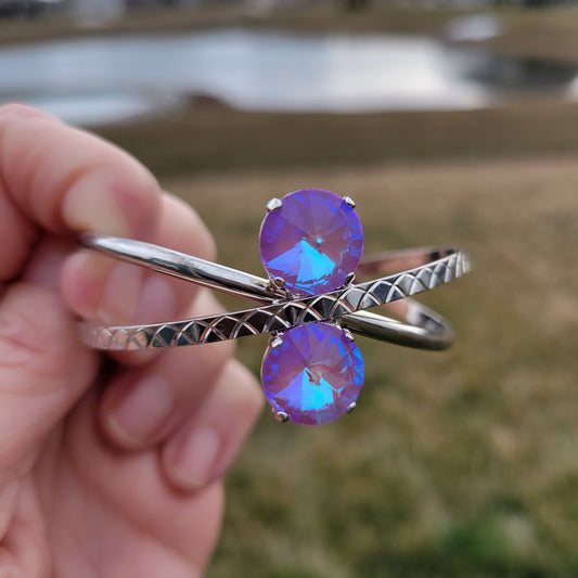 A hand holding a silver-toned wrap cuff bracelet with two round iridescent purple-blue crystals, set against a blurred outdoor background.