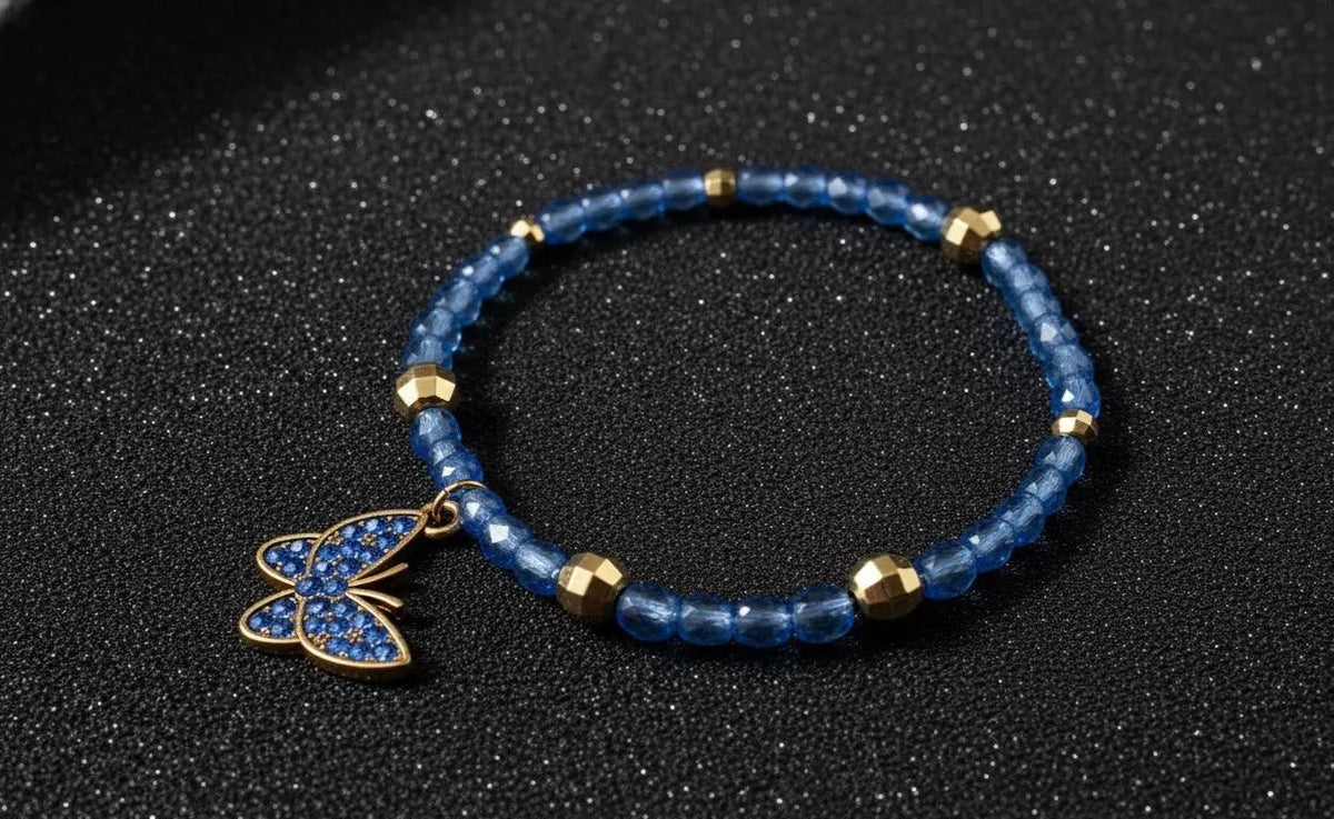 A beaded stretch bracelet featuring translucent blue beads, small gold spacer beads, and a butterfly charm embellished with blue stones and gold trim, displayed on a dark textured background.