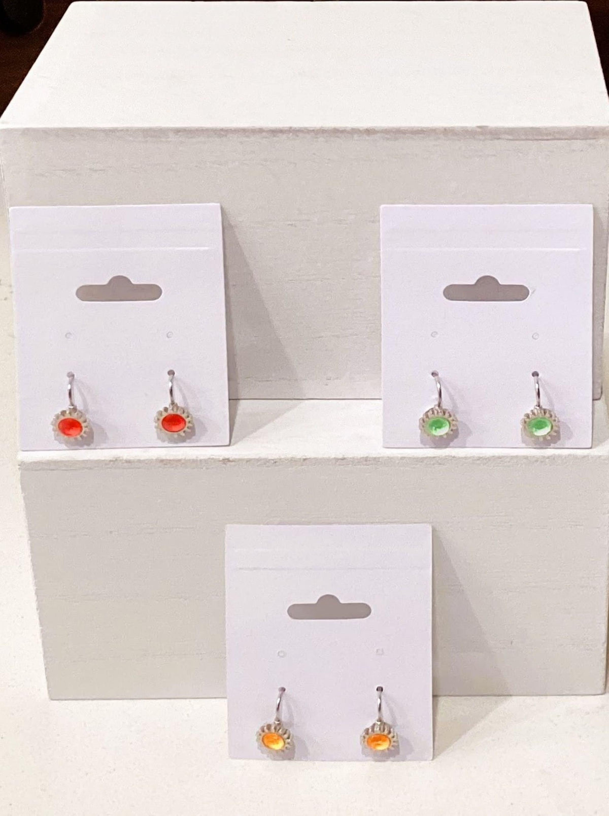 Three small dangling earrings displayed on white hanger cards against a white block background. The left pair has orange stones, the right top pair has green stones, and the bottom pair has yellow-orange stones, all with silver-toned settings.