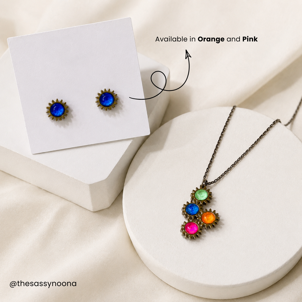 Jewelry display showing a matching necklace and stud earrings on white display blocks. The pieces have blue gemstone centers surrounded by bronze-toned petals, with the necklace also featuring green, orange, and pink stones.