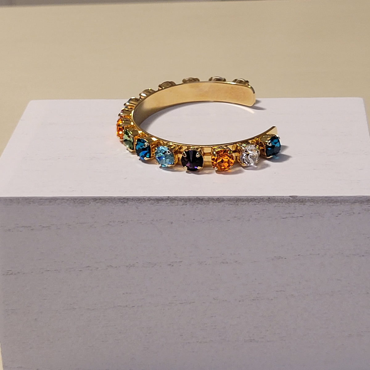 A gold-toned open cuff bracelet adorned with a row of faceted gemstones in blue, orange, black, and clear, displayed on a white block against a beige background.