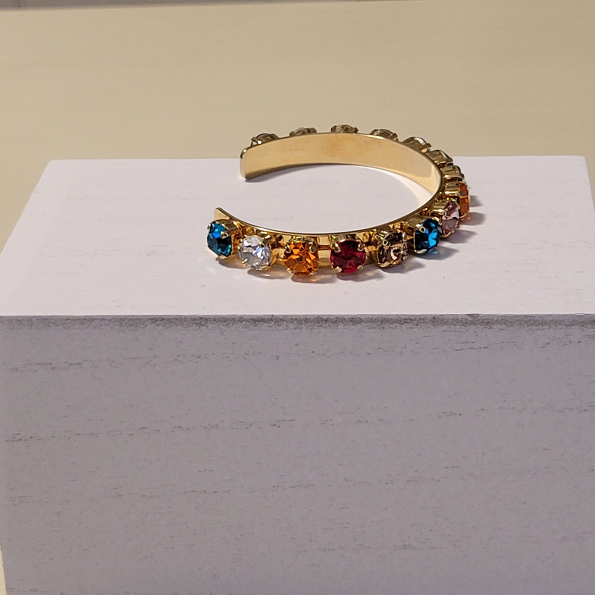 A gold-toned cuff bracelet with a row of colorful faceted rhinestones in blue, white, orange, and red, displayed on a white block against a beige background.