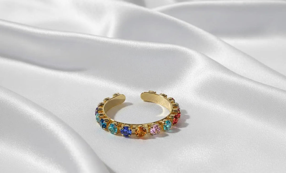 A gold cuff bracelet adorned with a row of colorful gemstones, resting on draped white satin fabric.