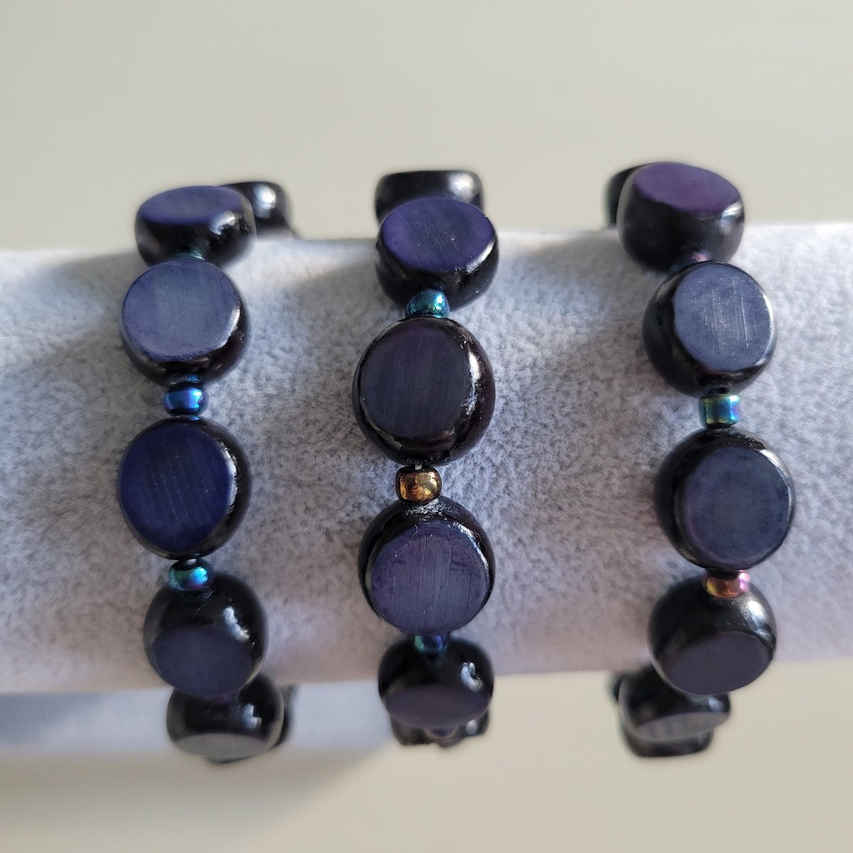 Three beaded stretch bracelets with dark round beads and small iridescent spacer beads, displayed on a white ring stand.