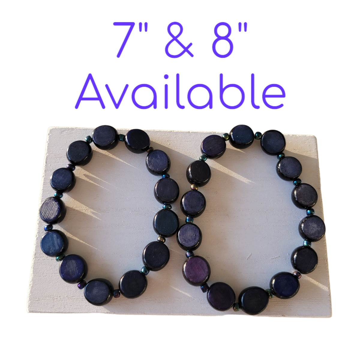 Two beaded bracelets displayed on a white rectangular board against a white background, with purple text above reading '7" & 8" Available'. The bracelets are made of dark round beads with a blue-purple iridescent sheen.