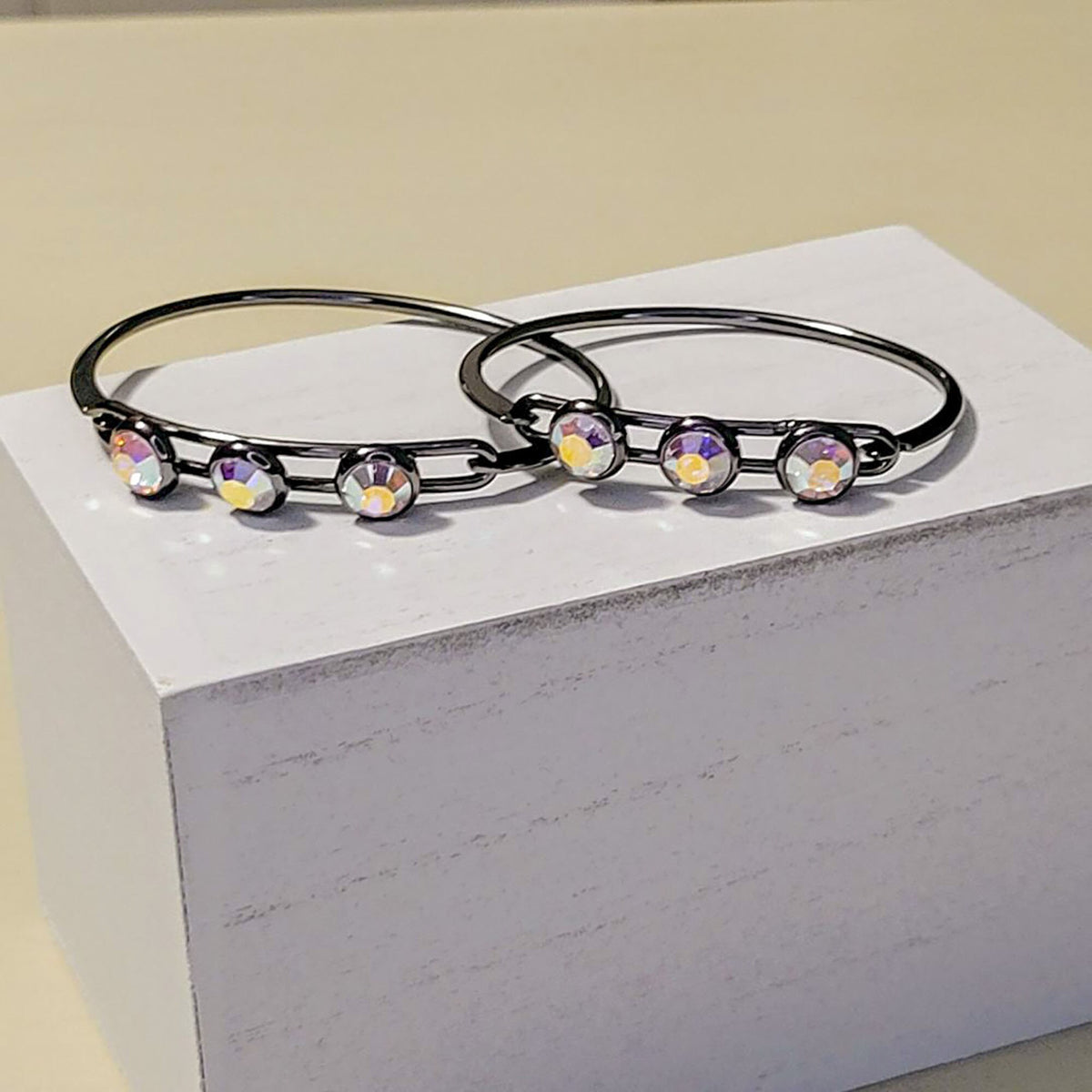 Two slim black metal bangles with five iridescent crystal charms each, displayed on a white block against a light beige background.